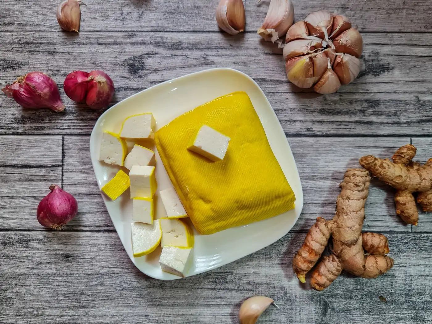 Fresh turmeric root, ginger, garlic and shallots around a plate of turmeric stained tofu, the same aromatics that go into a Balinese spice paste