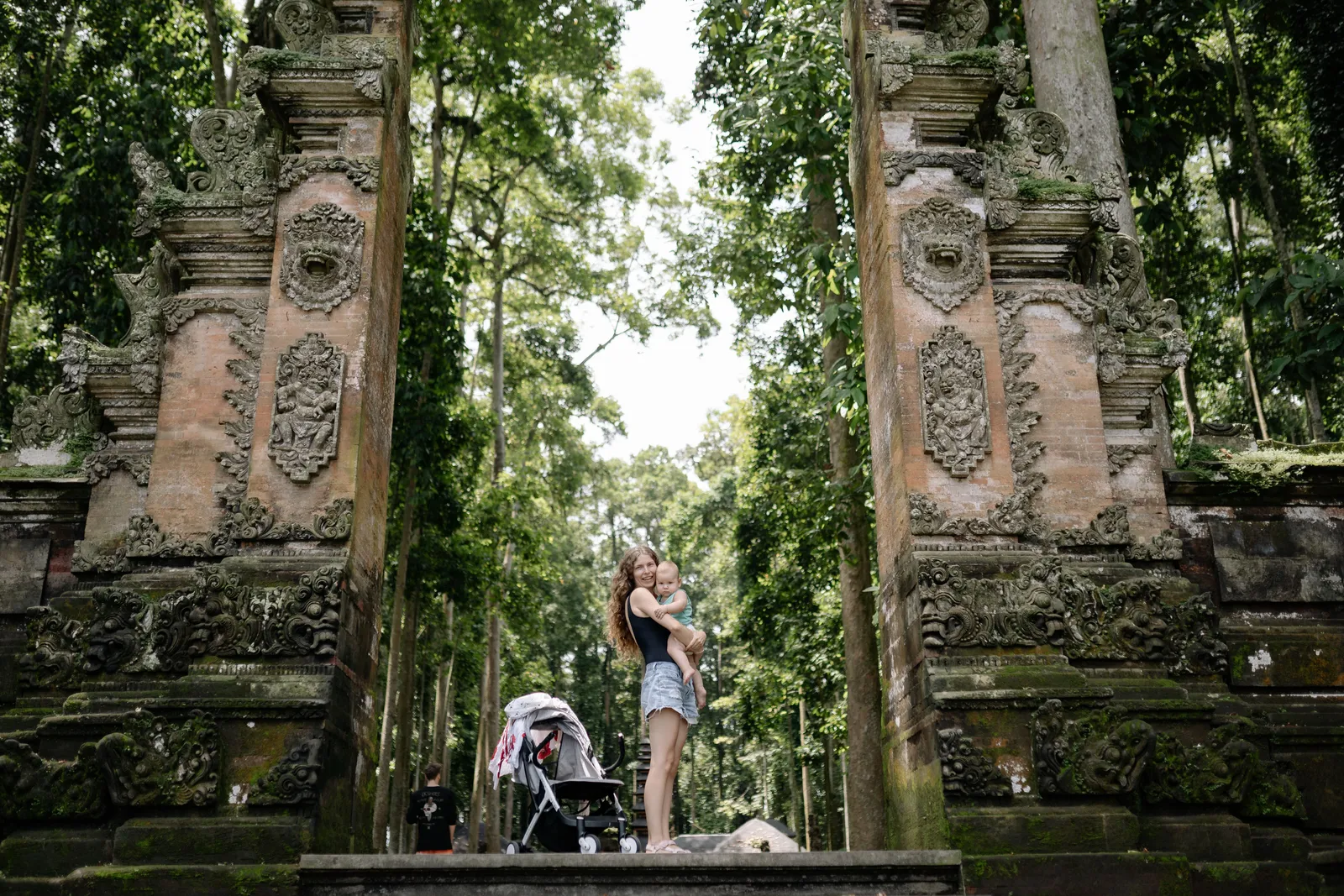A carved stone Balinese temple gate with visitors standing in the opening and jungle greenery behind