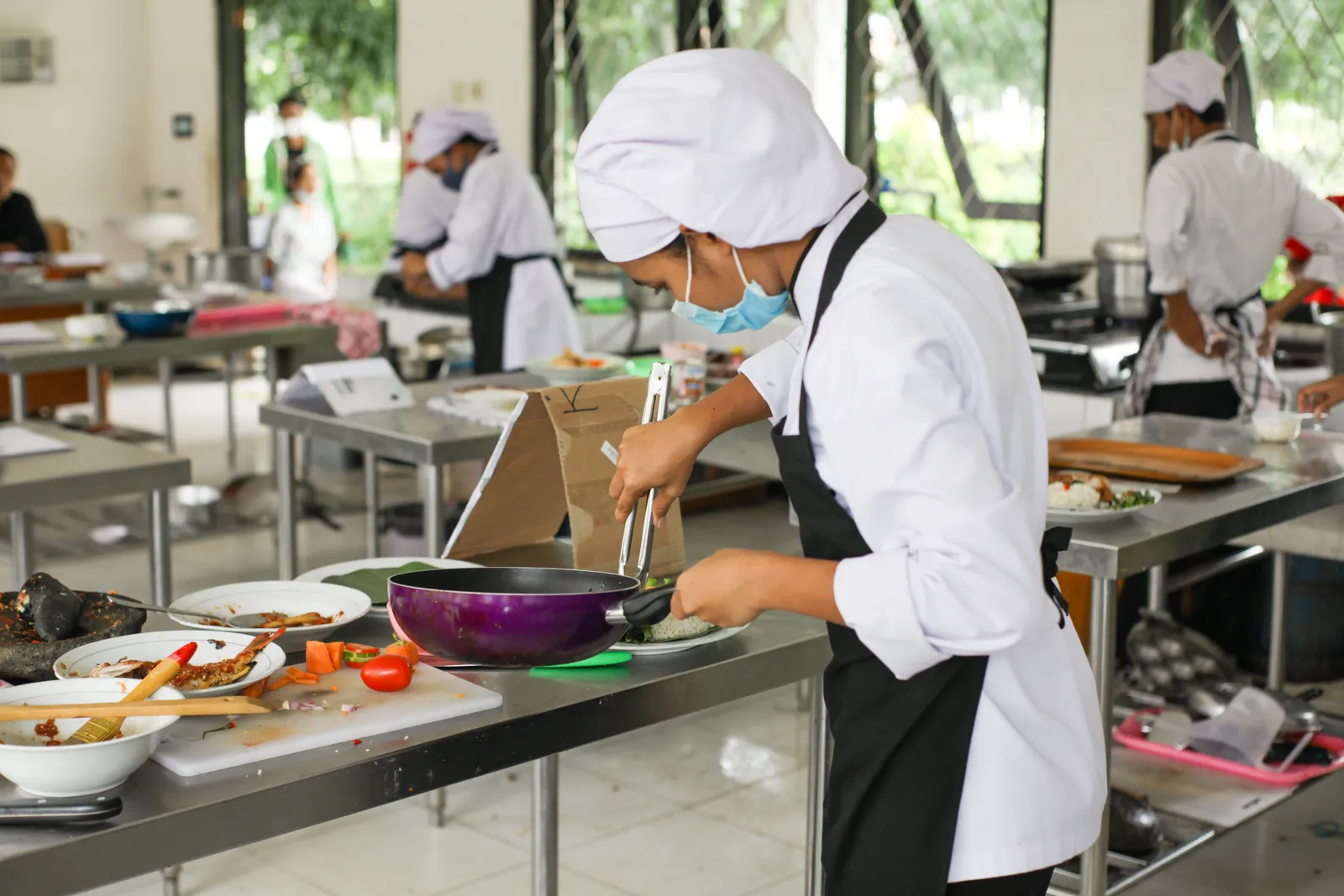 A chef in white uniform holding tongs in an open-air kitchen at a Balinese cooking class, Indonesia