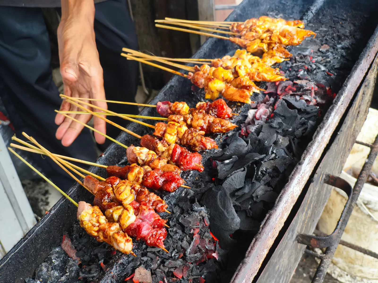 A cook grilling chicken satay over coals at a roadside food stall in Bali