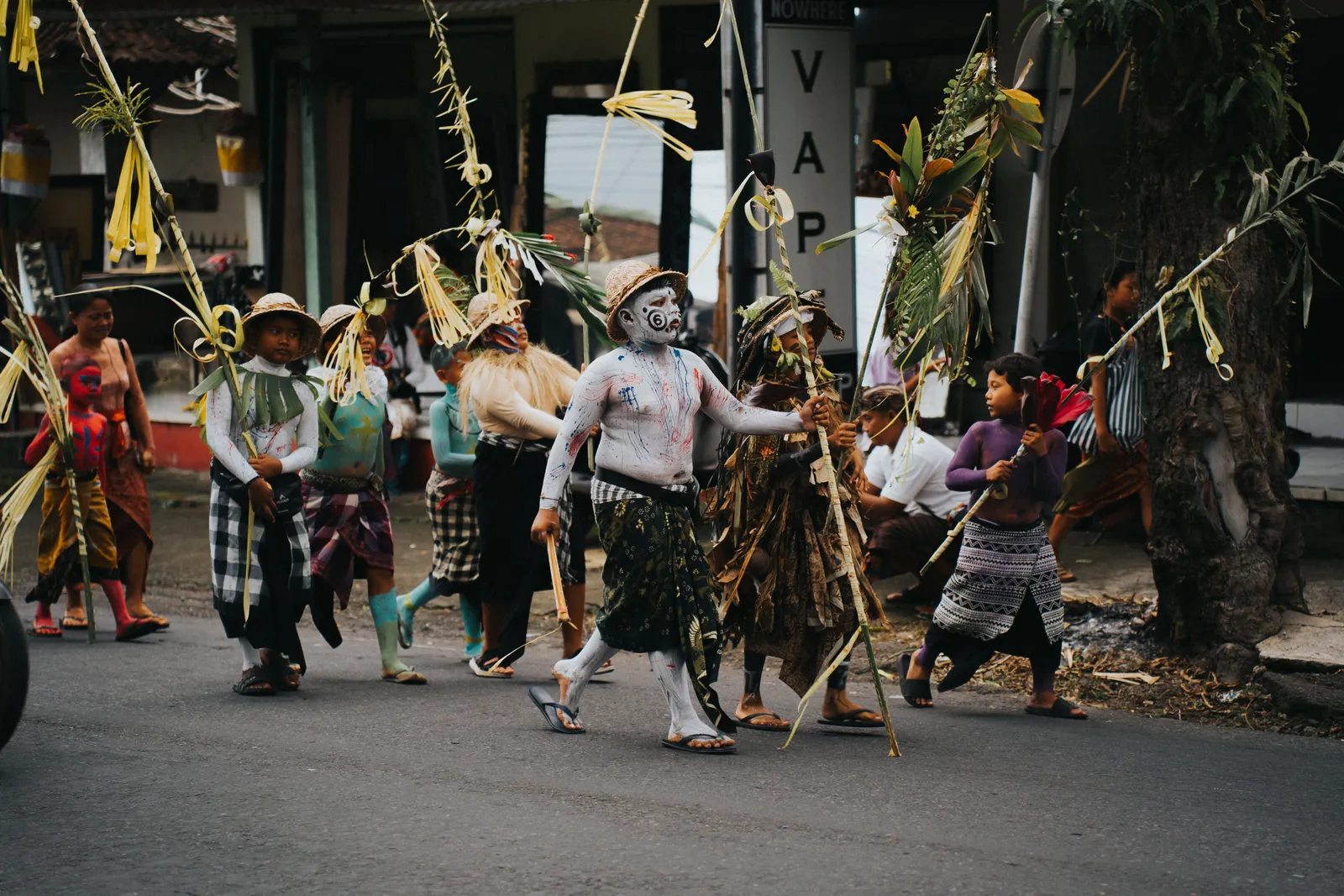A Balinese village procession with costumed participants carrying tall decorated bamboo poles through the street