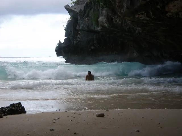 A sandy cove beneath a tall dark limestone cliff, white surf breaking over the rocks