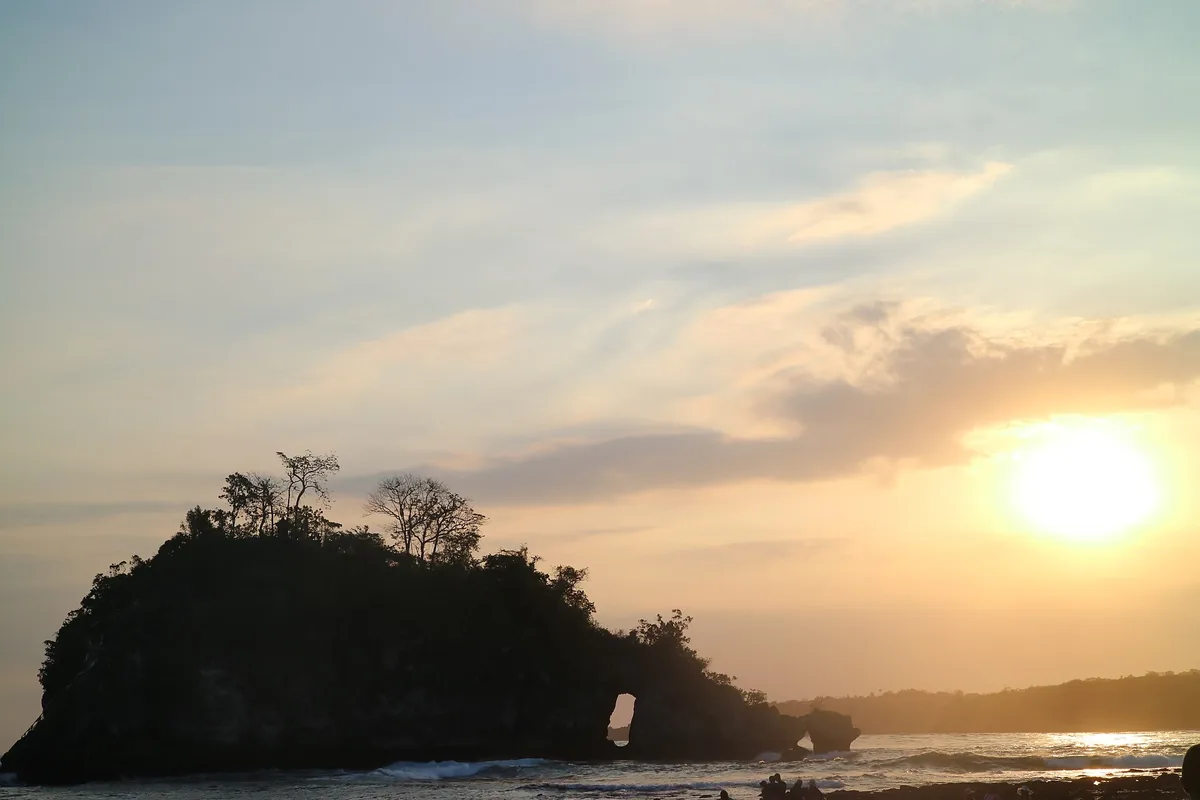 Sunset over a calm bay, a dark wooded headland at one side and low waves on pale sand