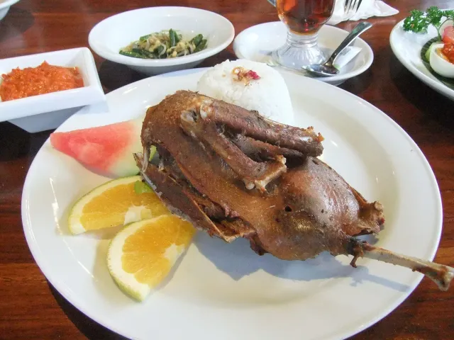 A crispy fried half duck on a plate with white rice, sambal and orange slices