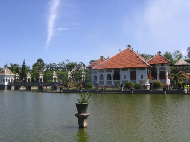 A white pavilion with a red tiled roof across a wide pond, a stone planter standing in the water