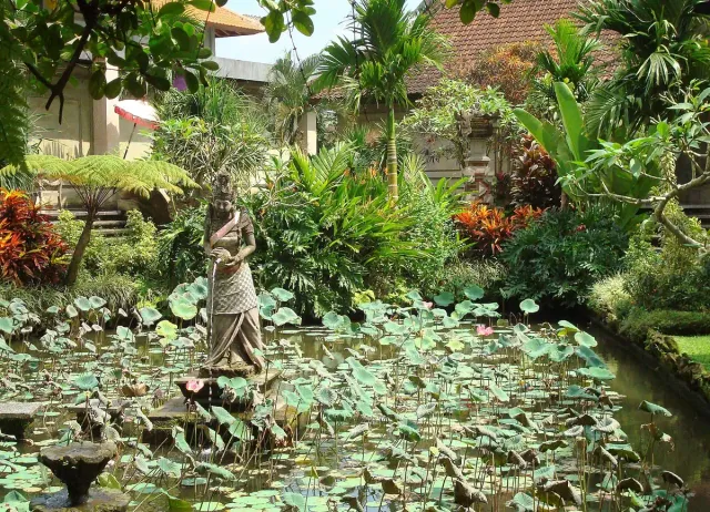 A lotus pond with a stone statue at its centre, a tropical garden and traditional buildings behind
