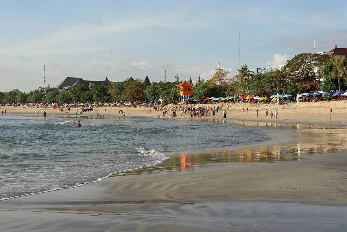 A wide busy beach with bathers, a palm treeline and parasols in afternoon light
