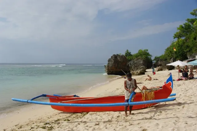 A red traditional outrigger boat on white sand, turquoise water and limestone rocks at the end of the cove