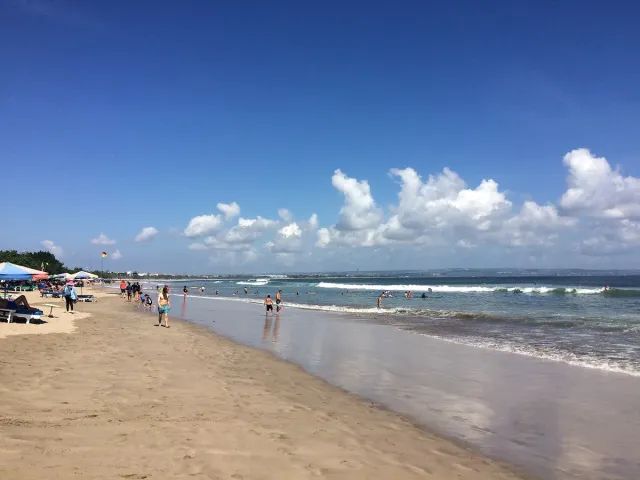 A wide strip of sand with umbrellas and sun loungers, low waves and people in the shallows
