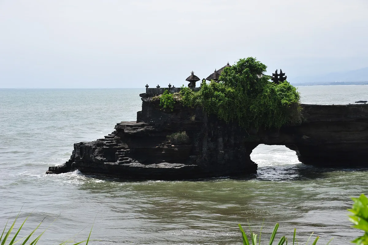 A dark sea rock with a natural arch and shrine roofs wrapped in greenery above it