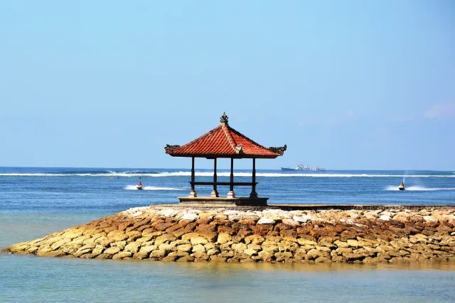 A pavilion with a red tiled roof on a stone breakwater, jet skis crossing the water behind