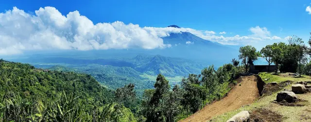 A green ridge with a dirt path, and above it a large volcano rising over a layer of cloud