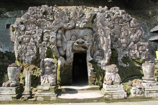 A cave mouth carved into rock and framed by stone reliefs of a face and creatures, with statues on either side