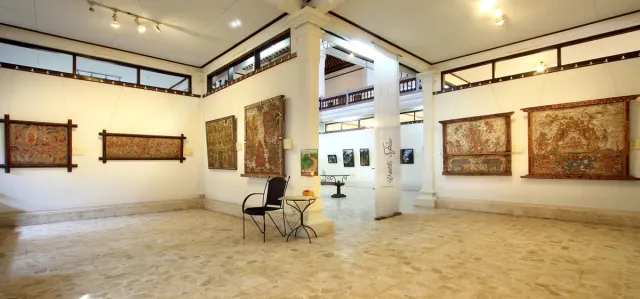 A bright gallery hall with framed paintings on the walls, a white column and chairs at its centre