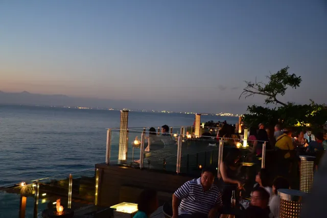 Bar terraces on a cliff edge above the sea at dusk, with lit lanterns and guests at the tables
